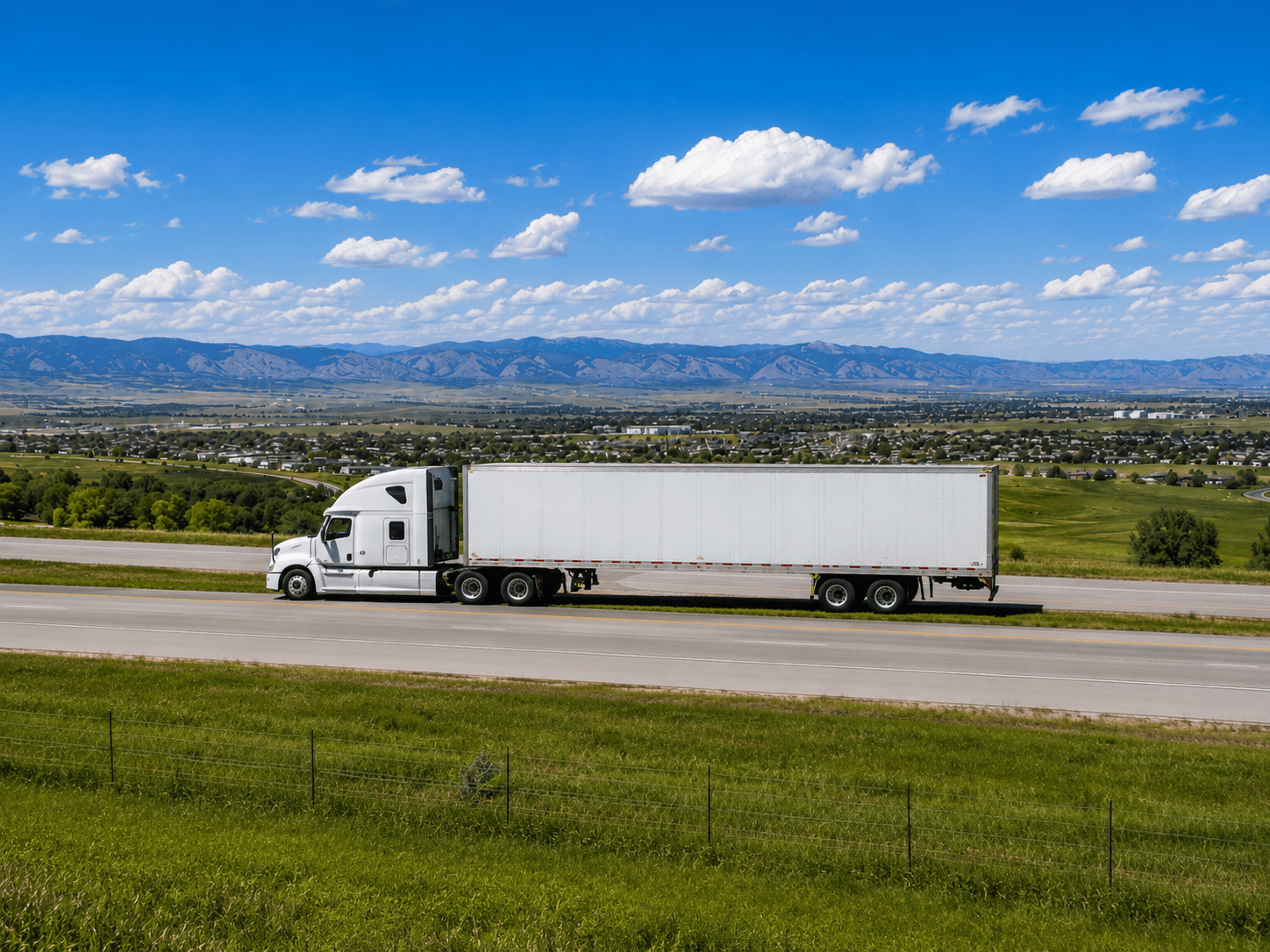 White semi truck hauling a dry van trailer along a highway with open green fields, scattered homes, and mountain views under a bright blue sky.