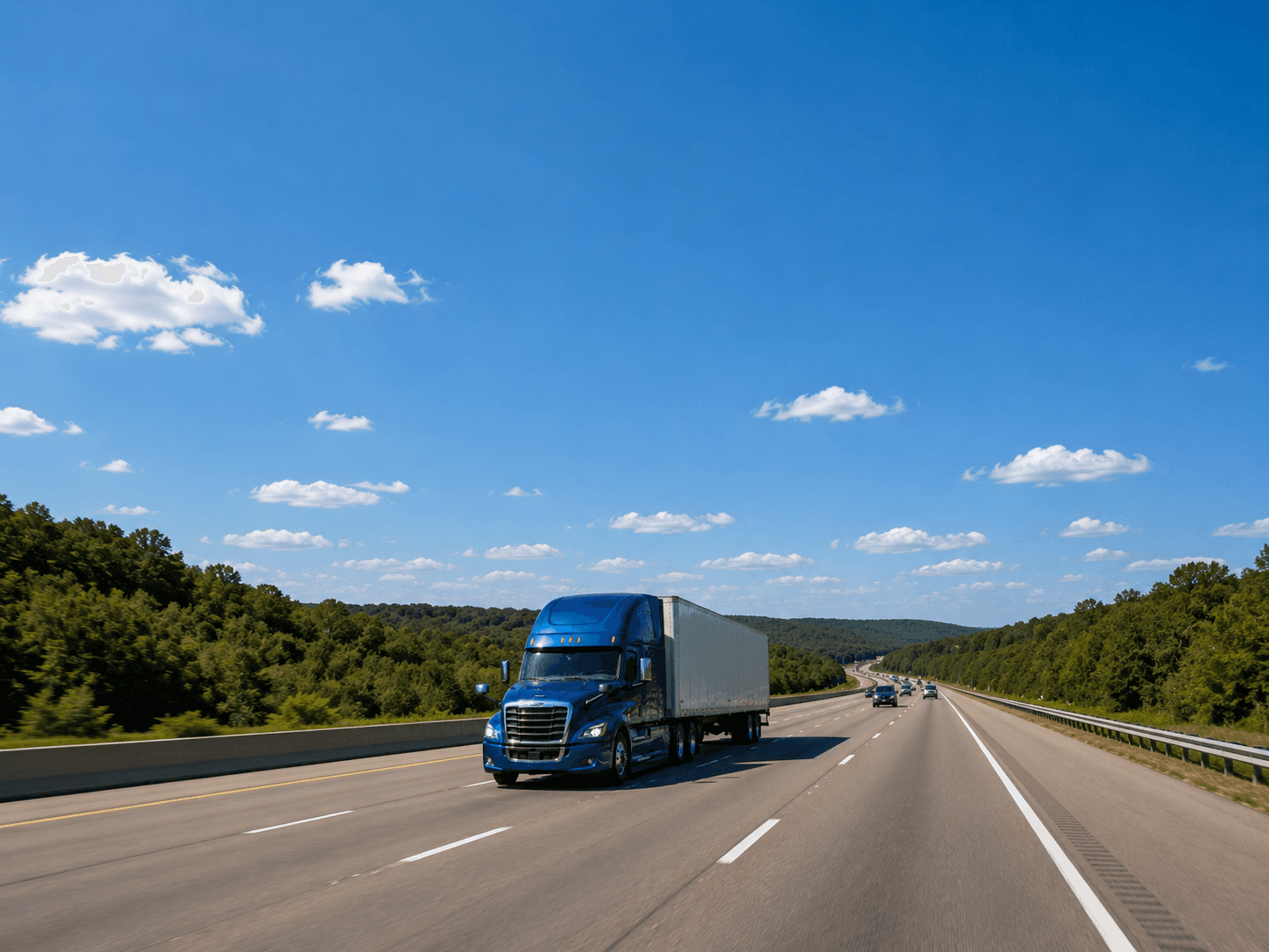 A blue semi-truck driving on a highway surrounded by green trees under a bright blue sky with scattered clouds.